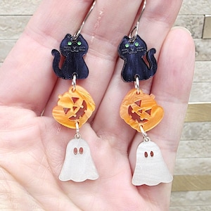 May include: A pair of Halloween-themed dangle earrings. Each earring features a black cat, an orange jack-o'-lantern, and a white ghost charm. The earrings are suspended from silver-colored ear wires, creating a festive accessory.