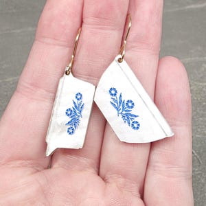 May include: White and silver earrings with a blue floral design. The earrings are made from repurposed china and feature gold-colored ear wires. The earrings are held in a hand.