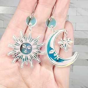 May include: A pair of celestial-themed earrings. One earring features a sun with a smiling face, and the other has a crescent moon with a star pendant. Both earrings have a teal and silver color scheme and dangle from a small, round stud.