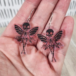 May include: A pair of pink glitter skeleton fairy earrings with black wings and silver hooks.