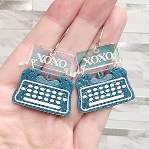 May include: A pair of dangle earrings shaped like vintage typewriters in a teal speckled finish with white keys. Above the typewriter body, a clear rectangular piece features the text XOXO in white. The earrings hang from silver-toned hooks.