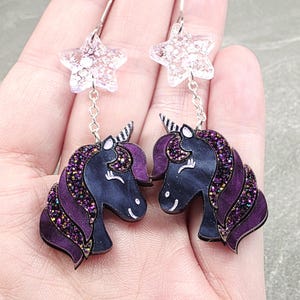 May include: A pair of whimsical earrings featuring a unicorn design. The earrings have a dark blue body with purple glitter accents and a purple mane. Each earring is connected to a clear star-shaped charm with silver hooks.