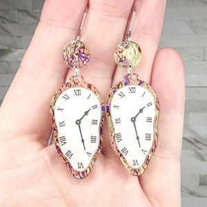 May include: A pair of unique earrings featuring a melting clock design. The white clock faces have black hands and Roman numerals. The earrings are suspended from silver-colored hooks and have iridescent, circular tops with purple and gold accents.