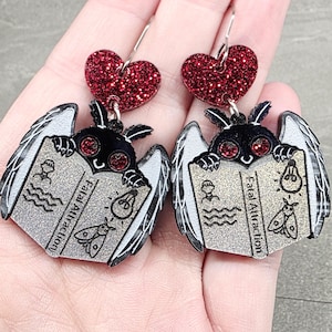 May include: A pair of dangle earrings featuring a black moth with red eyes reading a silver book titled Fatal Attraction. The moth has white wings and is topped with a red glitter heart. The book includes small black illustrations of a person, a lightbulb, and a moth.