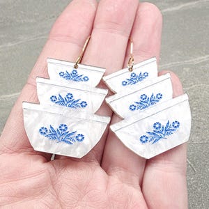 May include: A pair of earrings featuring a stacked bowl design. Each bowl is white with a blue floral pattern. The earrings are held by a person's hand, and the hoops are gold.