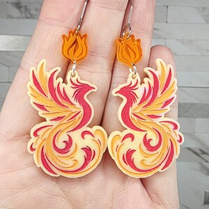 May include: A pair of dangle earrings featuring a phoenix design in shades of cream, yellow, and red. Each earring has a small orange flame-shaped topper attached to a silver-toned metal hook. The phoenix figures are layered with intricate cutouts.