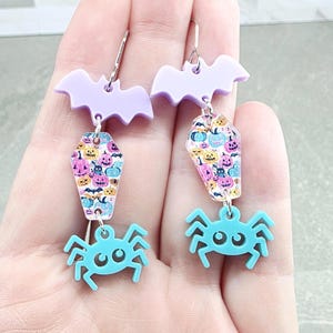 May include: Dangle earrings featuring a light purple bat at the top, a central coffin-shaped charm with a colorful pattern of pumpkins and ghosts, and a bright turquoise spider at the bottom, all connected by silver-toned metal jump rings.