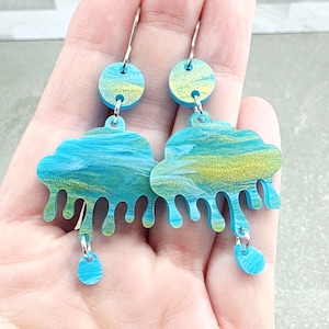 May include: A pair of dangle earrings featuring a cloud-shaped charm with a marbled blue, teal, and gold finish. Each earring has a small circular stud at the top and a single teardrop-shaped pendant hanging from the bottom of the cloud.