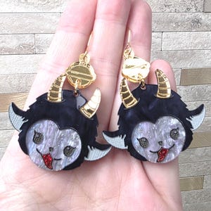 May include: A pair of dangle earrings featuring a black and iridescent white creature face with gold-toned horns and a red glitter tongue. The earrings are attached to gold-toned circular studs and feature a detailed, layered acrylic design.