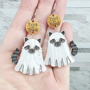May include: Dangle earrings featuring white raccoon ghosts wearing sheets with striped tails. Above each ghost is a round, gold glitter charm with the black text Trick or Trash. The earrings have silver-toned metal hooks for attachment.