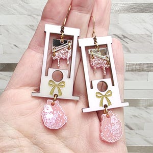 May include: A pair of dangle earrings featuring a white guillotine frame with a gold glitter bow accent. The guillotine blade is silver with pink glitter drips, and a pink floral-patterned head charm hangs from the bottom of each earring.