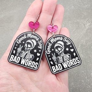 May include: A pair of dangle earrings featuring a black arch-shaped pendant with a white skeleton graphic making a peace sign. The text reads Good Moms Say BAD WORDS. Above the pendant is a small pink heart-shaped connector attached to a silver hook.
