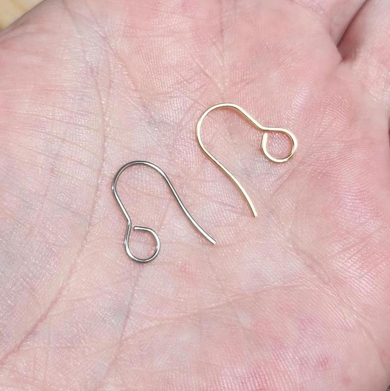 Op de afbeelding: Twee oorhaken, een zilveren en een gouden, worden getoond op een hand. De haken zijn beide open en klaar voor gebruik.