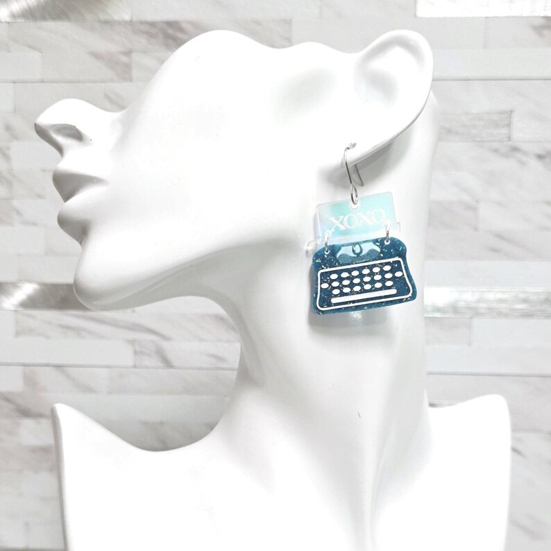 May include: A dangling earring featuring a dark blue glittery typewriter charm with white keys and a light blue rectangular top piece displaying the white text XOXO, suspended from a silver-toned metal hook on a white display bust.
