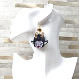 May include: A dangling earring featuring a dark purple and black creature head with two gold-toned horns, a light purple face, and a small red accent on the mouth. The charm hangs from a gold-toned metal hook and a small circular gold-toned connector.
