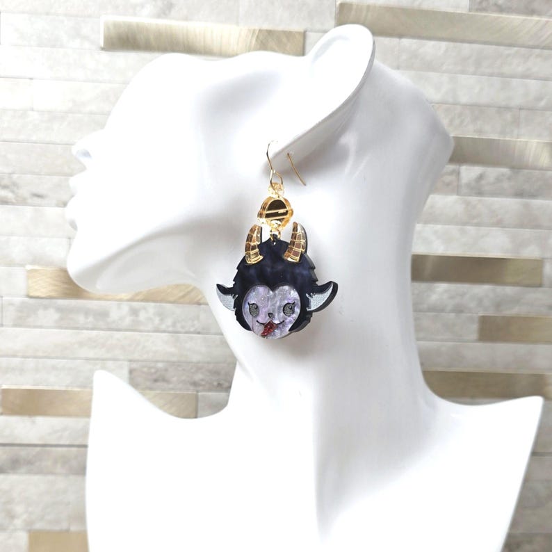 May include: A dangling earring featuring a dark purple and black creature head with two gold-toned horns, a light purple face, and a small red accent on the mouth. The charm hangs from a gold-toned metal hook and a small circular gold-toned connector.