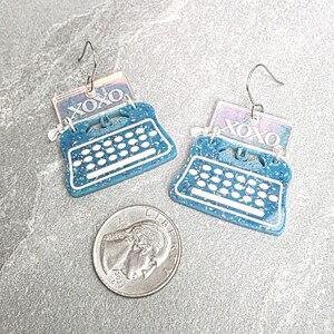 May include: A pair of teal typewriter-shaped earrings with white keys and a rectangular top piece featuring the text XOXO in white. The earrings have silver-toned metal hooks and are shown next to a United States five-cent coin for scale.