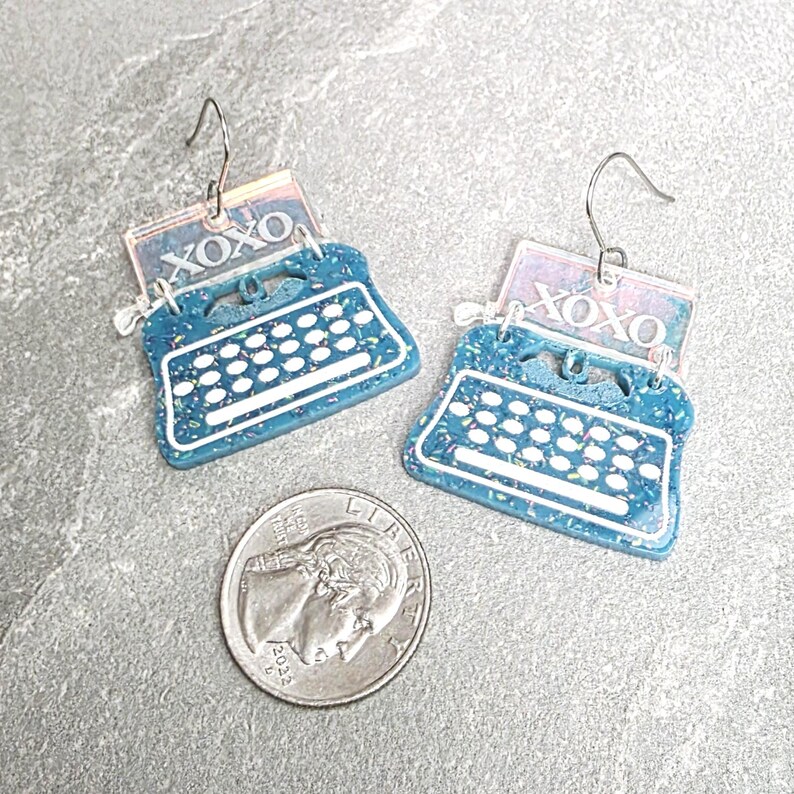 May include: A pair of teal typewriter-shaped earrings with white keys and a rectangular top piece featuring the text XOXO in white. The earrings have silver-toned metal hooks and are shown next to a United States five-cent coin for scale.