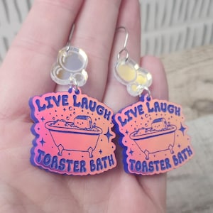 May include: A pair of iridescent pink and blue earrings with a toaster in a bathtub design. The earrings feature the text "Live Laugh Toaster Bath".