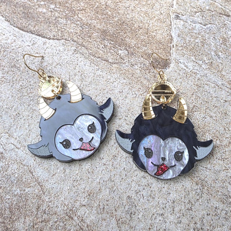 May include: A pair of dangle earrings featuring a stylized creature head with black hair, iridescent white face, gold-toned horns, and a red tongue. The earrings include gold-toned metal hooks and circular connectors, showcasing a detailed design.