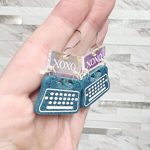 May include: Teal typewriter earrings with white keyboard details and a rectangular iridescent top piece featuring the text XOXO in white. The earrings hang from silver-toned metal hooks and have a speckled texture on the typewriter body.