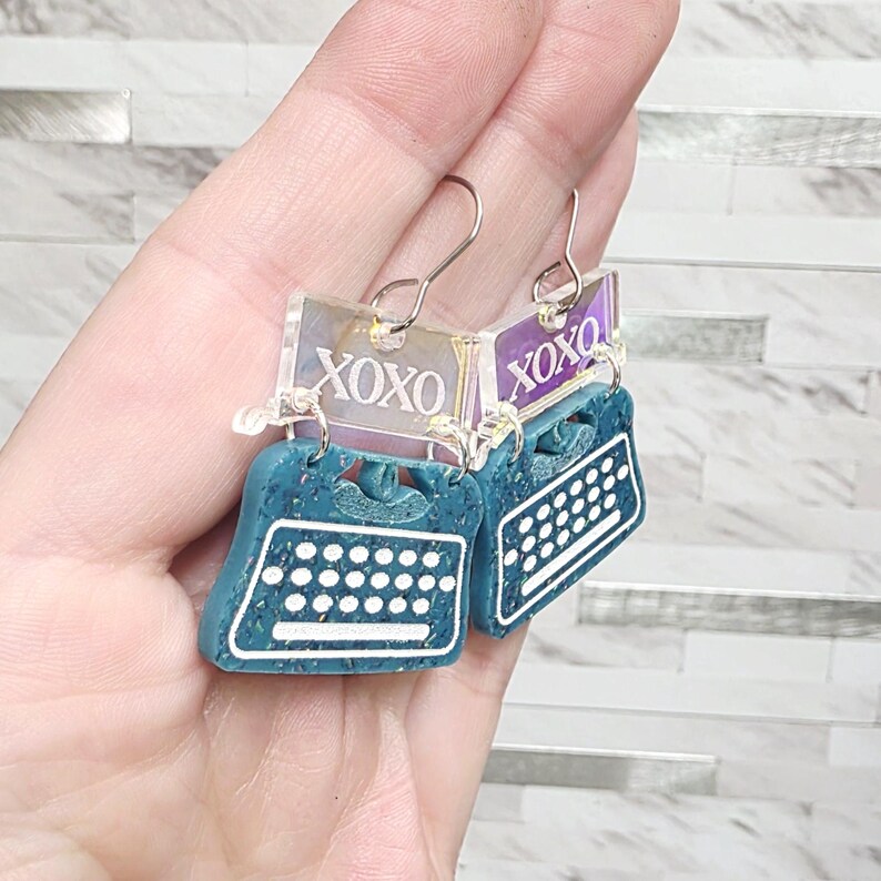 May include: Teal typewriter earrings with white keyboard details and a rectangular iridescent top piece featuring the text XOXO in white. The earrings hang from silver-toned metal hooks and have a speckled texture on the typewriter body.