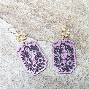 May include: A pair of glittery purple dangle earrings featuring a rectangular pendant with a black illustration of a person holding a cup, a bird on their shoulder, and flowers. The text reads RAGE... BUT MAKE IT Floral. Gold flower-shaped connectors.