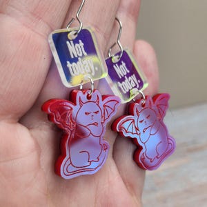 May include: A pair of earrings featuring a red devil design with iridescent wings and a matching tag that reads "Not today." The earrings have silver-colored hooks.