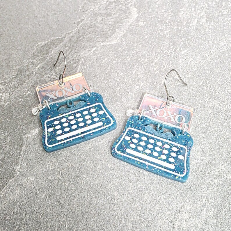 May include: A pair of dangle earrings featuring a teal typewriter charm with white keys and a speckled finish. Attached to the top of each typewriter is a rectangular, iridescent clear acrylic piece with the text XOXO printed in white.