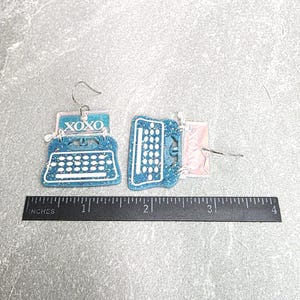 May include: A pair of blue glitter typewriter earrings with white keys and the text XOXO printed on the paper section. The earrings are positioned above a black ruler showing a width of approximately 3.5 inches for the pair.