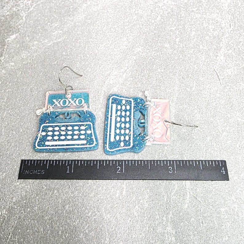 May include: A pair of blue glitter typewriter earrings with white keys and the text XOXO printed on the paper section. The earrings are positioned above a black ruler showing a width of approximately 3.5 inches for the pair.