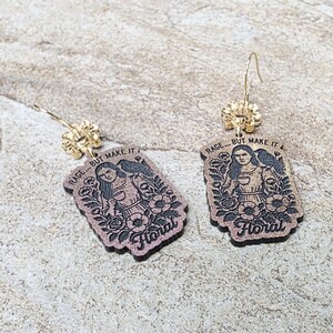 May include: A pair of dangling earrings featuring rectangular wooden pendants with a dark brown finish. Each pendant displays an engraved illustration of a person surrounded by flowers and the text RAGE... BUT MAKE IT Floral.