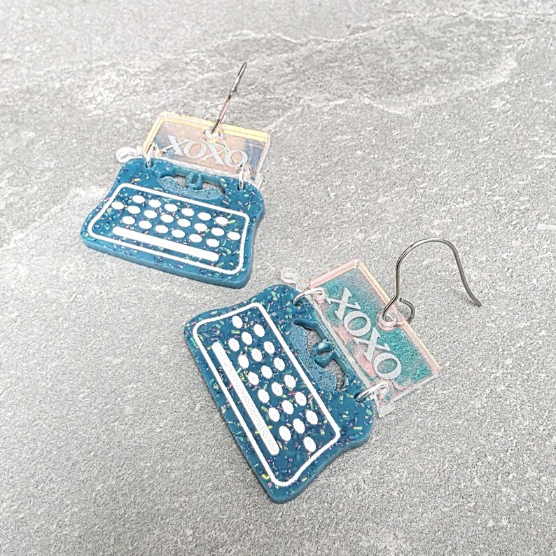May include: A pair of dangle earrings featuring a teal typewriter design with white keys and a clear rectangular topper engraved with the text XOXO. The typewriter body has a speckled texture and silver-toned metal earring hooks.