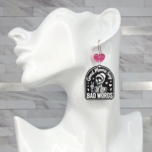 May include: A dangling earring featuring a small pink heart charm attached to a larger black and white arched pendant. The pendant displays a skeleton graphic making a peace sign with the text Good Moms Say BAD WORDS in white lettering.