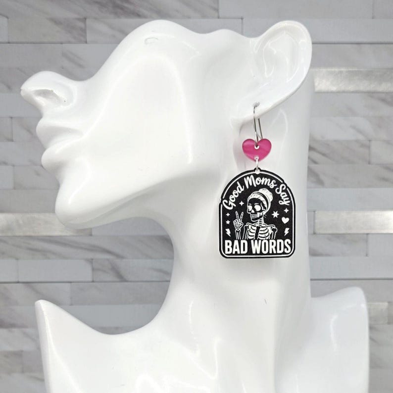 May include: A dangling earring featuring a small pink heart charm attached to a larger black and white arched pendant. The pendant displays a skeleton graphic making a peace sign with the text Good Moms Say BAD WORDS in white lettering.