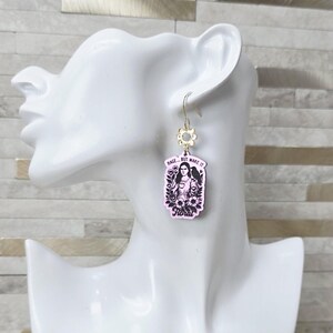 May include: A dangling earring featuring a rectangular light pink pendant with a black illustration of a person holding a cup, surrounded by flowers and a bird. The text reads RAGE... BUT MAKE IT Floral. The pendant hangs from a gold-toned flower charm.