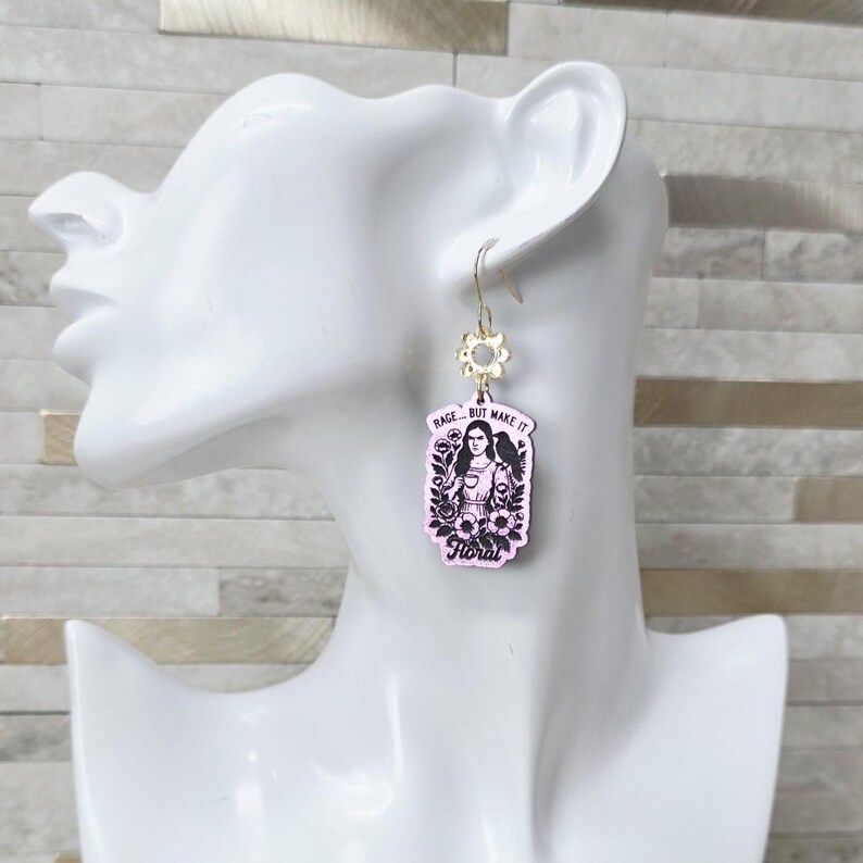 May include: A dangling earring featuring a rectangular light pink pendant with a black illustration of a person holding a cup, surrounded by flowers and a bird. The text reads RAGE... BUT MAKE IT Floral. The pendant hangs from a gold-toned flower charm.