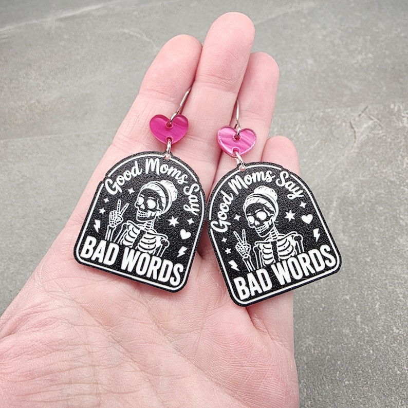 May include: A pair of dangle earrings featuring a black arch-shaped pendant with a white skeleton graphic making a peace sign. The text reads Good Moms Say BAD WORDS. Above the pendant is a small pink heart-shaped connector attached to a silver hook.
