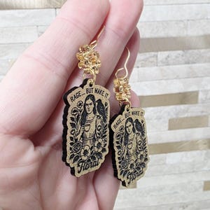 May include: A pair of dangle earrings featuring rectangular wooden pendants with a black engraved illustration of a person holding a cup, a bird on their shoulder, and flowers. The text RAGE... BUT MAKE IT Floral is engraved on each piece.