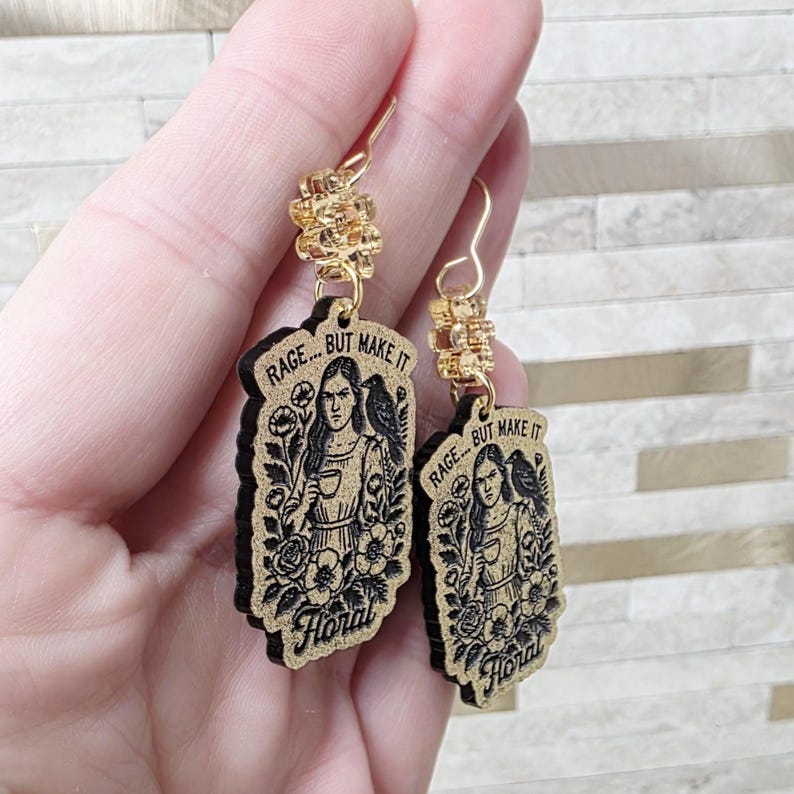 May include: A pair of dangle earrings featuring rectangular wooden pendants with a black engraved illustration of a person holding a cup, a bird on their shoulder, and flowers. The text RAGE... BUT MAKE IT Floral is engraved on each piece.