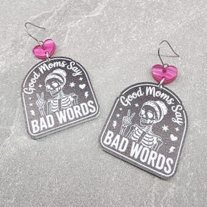 May include: A pair of dangle earrings featuring a black arch-shaped pendant with a white skeleton graphic, the text Good Moms Say BAD WORDS, and small stars and hearts. Each earring has a pink heart-shaped connector and a silver-toned metal hook.