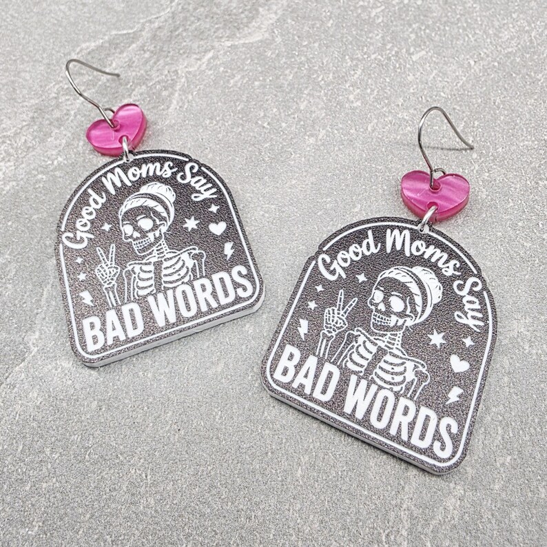 May include: A pair of dangle earrings featuring a black arch-shaped pendant with a white skeleton graphic, the text Good Moms Say BAD WORDS, and small stars and hearts. Each earring has a pink heart-shaped connector and a silver-toned metal hook.