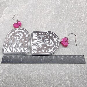 May include: A pair of dangle earrings featuring gray glittery arch-shaped pendants with a white skeleton graphic and the text "Good Moms Say BAD WORDS". Each earring has a pink heart-shaped accent and a metal hook, shown with a 4-inch ruler.