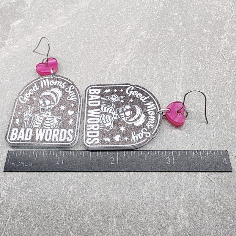 May include: A pair of dangle earrings featuring gray glittery arch-shaped pendants with a white skeleton graphic and the text "Good Moms Say BAD WORDS". Each earring has a pink heart-shaped accent and a metal hook, shown with a 4-inch ruler.