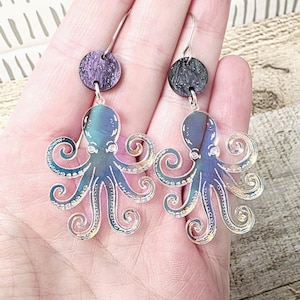 May include: A pair of dangle earrings featuring iridescent octopus charms with eight curled tentacles. Each octopus hangs from a small, circular stud with a textured, dark purple and black marbled finish, attached to silver-toned metal hooks.