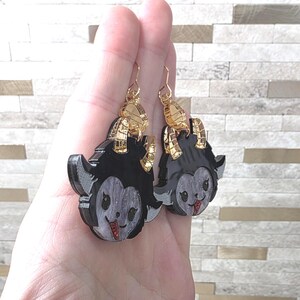 May include: A pair of dangle earrings featuring a black and grey glittery character head with horns and a red tongue. The earrings have gold-toned metal hooks and accents, showcasing a layered design with a glossy finish and detailed facial features.
