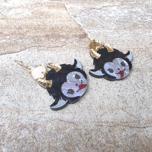 May include: A pair of dangle earrings featuring a black and iridescent white heart-shaped face with gold-toned horns and a red glitter tongue. The earrings are suspended from gold-toned metal hooks and display a stylized creature design.