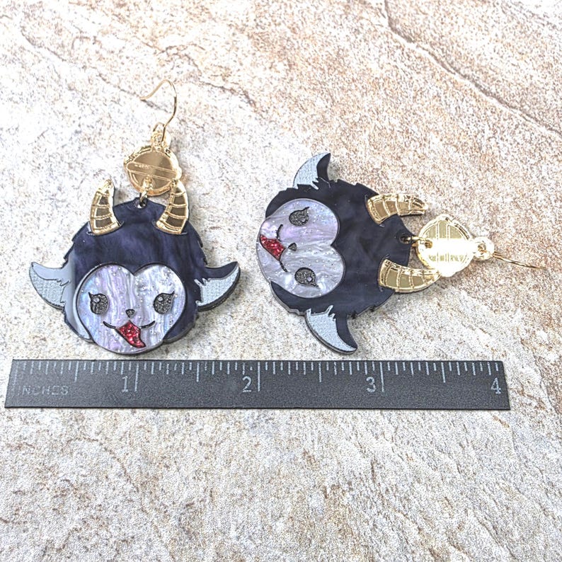 May include: A pair of dangle earrings featuring a dark purple and iridescent white creature face with gold-toned horns and a red tongue. The earrings are positioned above a black ruler, showing a width of approximately 3.5 inches for the pair.