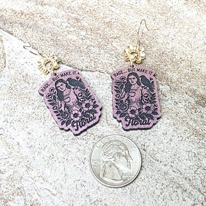May include: A pair of purple dangle earrings featuring a printed illustration of a person with a raven and flowers, with the text RAGE... BUT MAKE IT Floral. The earrings are shown next to a U.S. quarter coin for scale, highlighting the size.