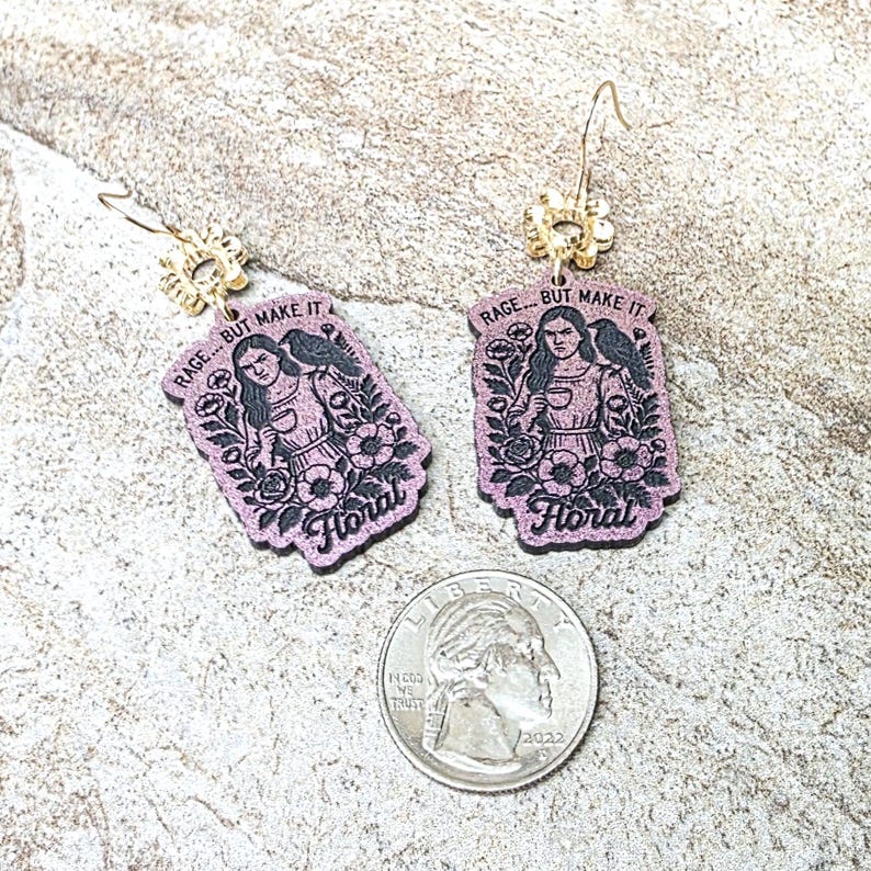 May include: A pair of purple dangle earrings featuring a printed illustration of a person with a raven and flowers, with the text RAGE... BUT MAKE IT Floral. The earrings are shown next to a U.S. quarter coin for scale, highlighting the size.